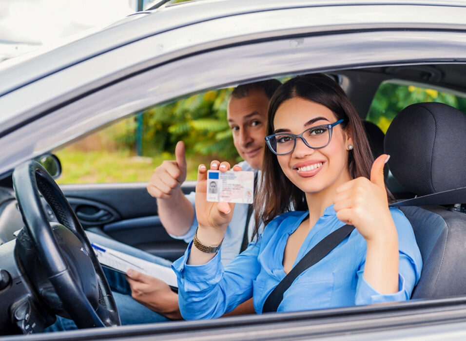 student giving the thumbs up, sitting in the car with her driving instructor.